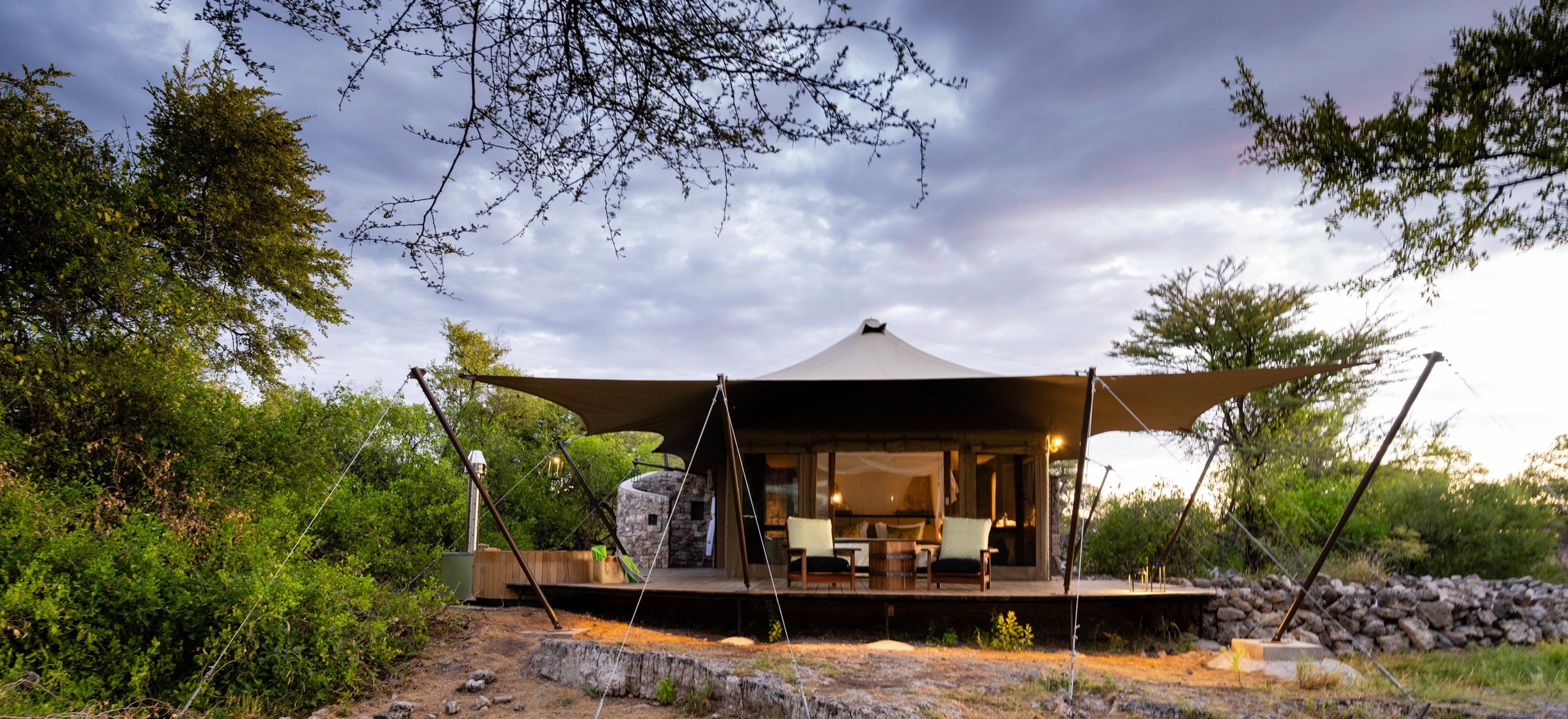A tented safari suite with chairs on a wooden deck surrounded by green bush