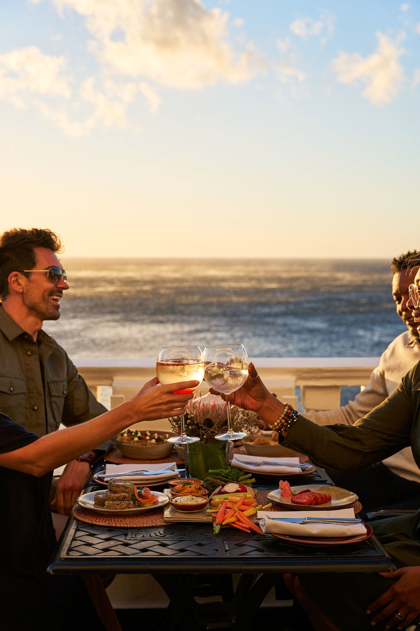 Travellers sharing a meal and raising their glasses beside the ocean at sunset