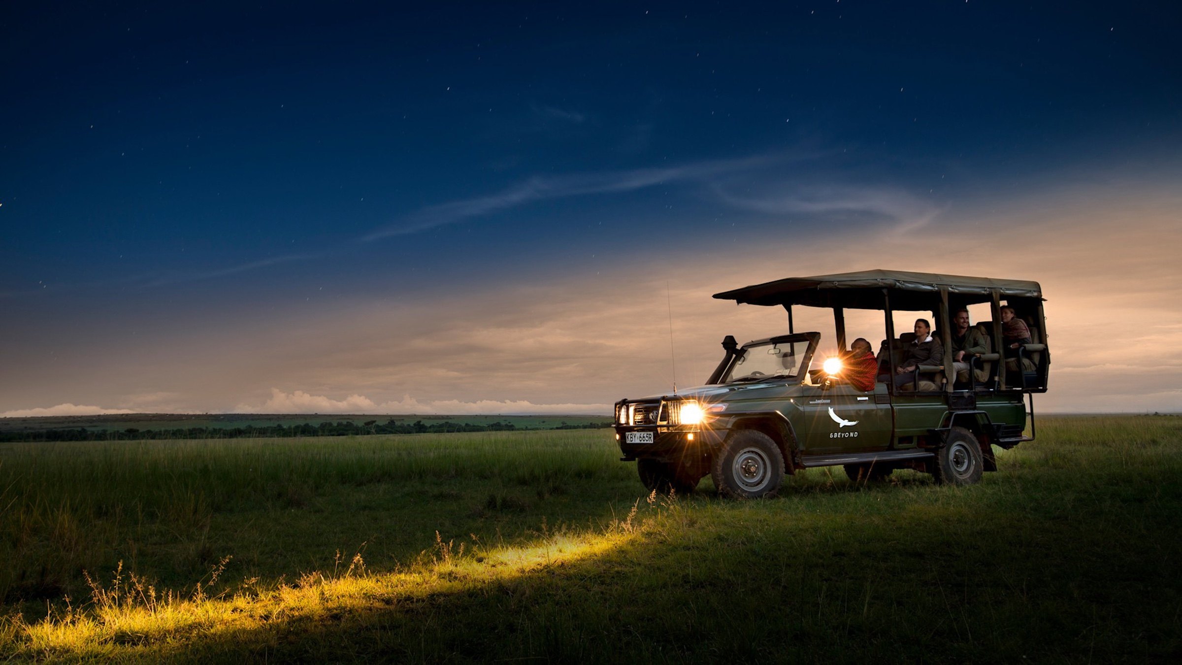 Guests in an open safari vehicle at dusk with its headlights illuminating the grass