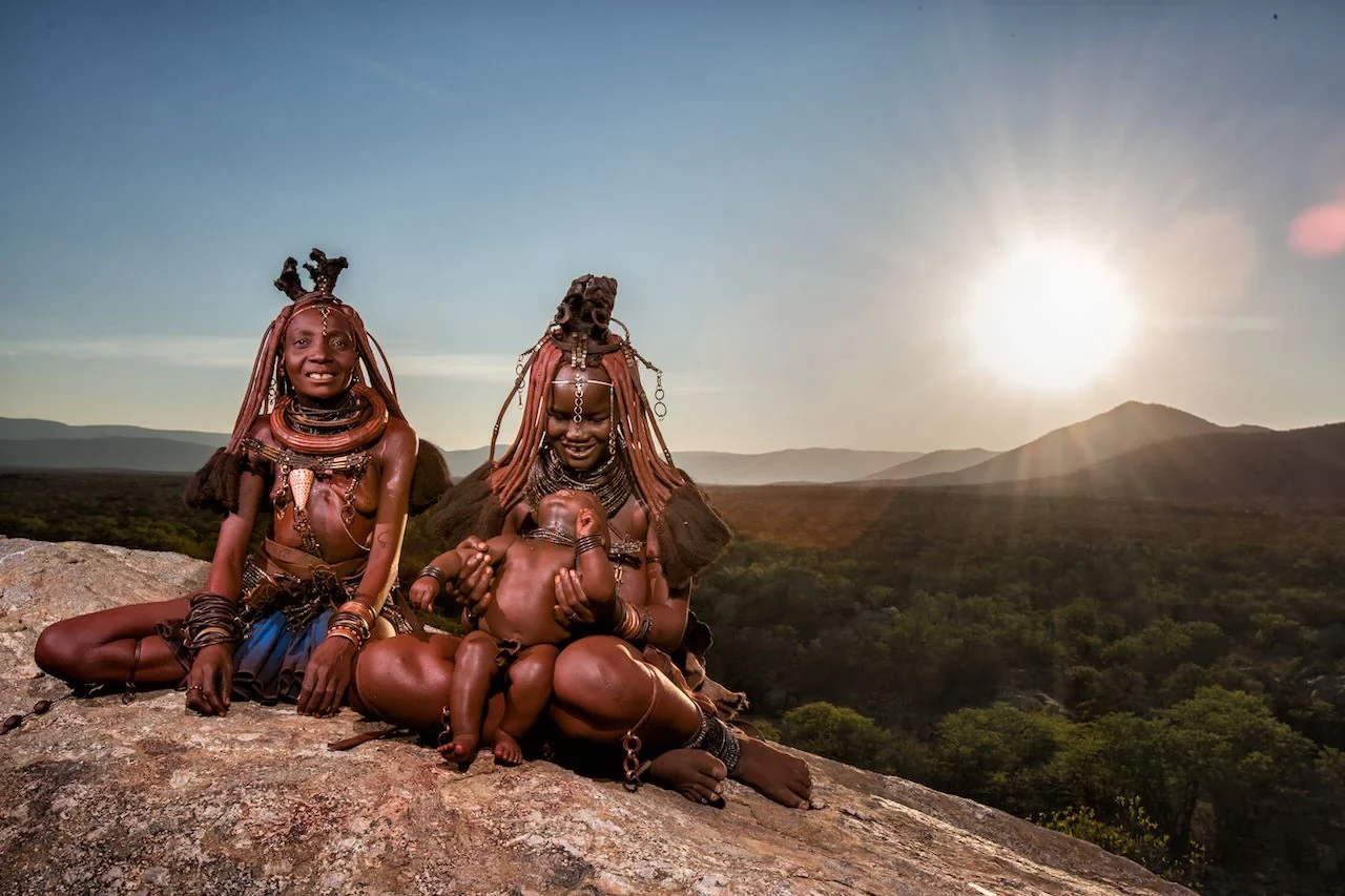 Two Himba women, one holding a child, seated on rocks in a mountain landscape