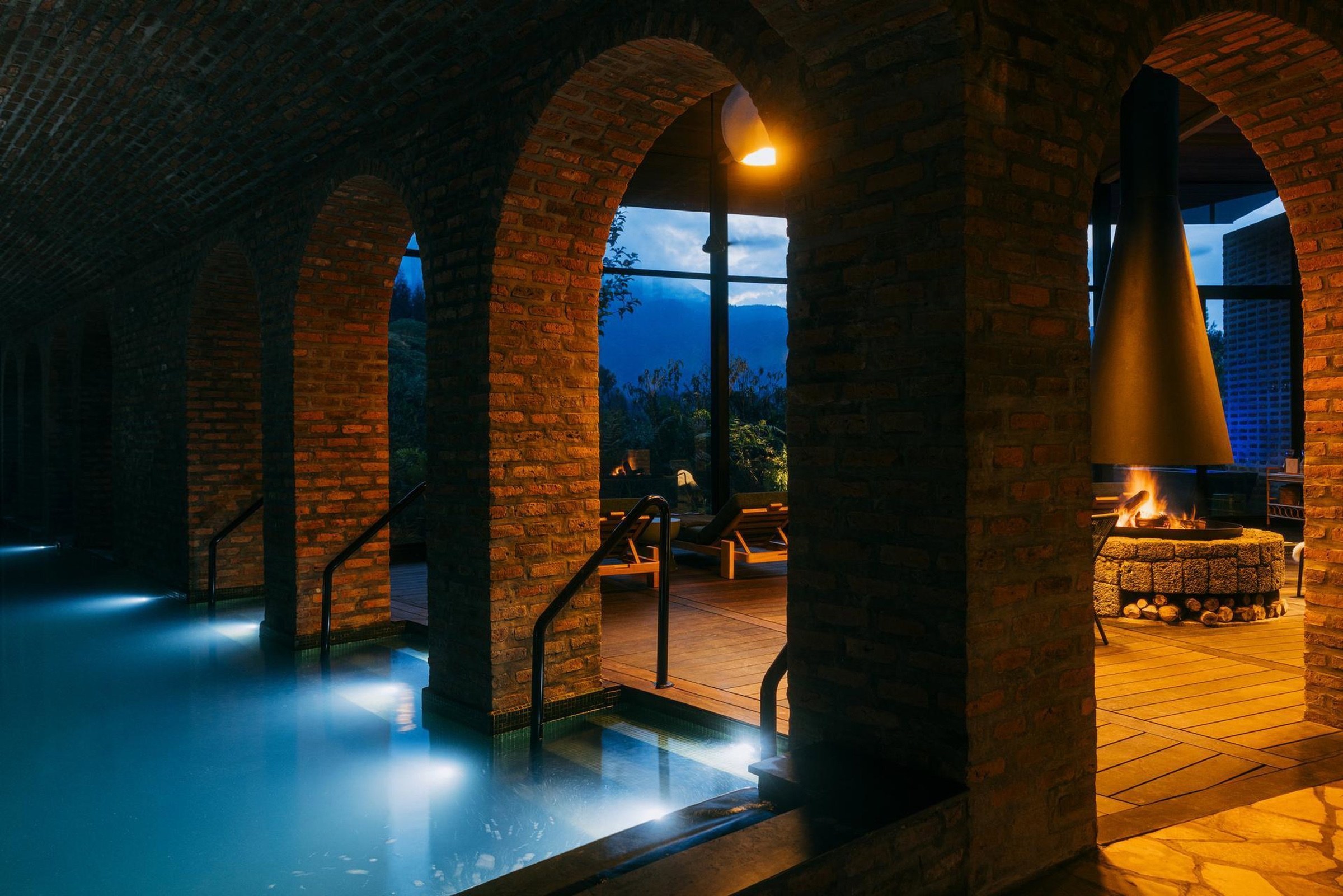 An illuminated lodge pool beside brick arches and a fireplace in Rwanda.
