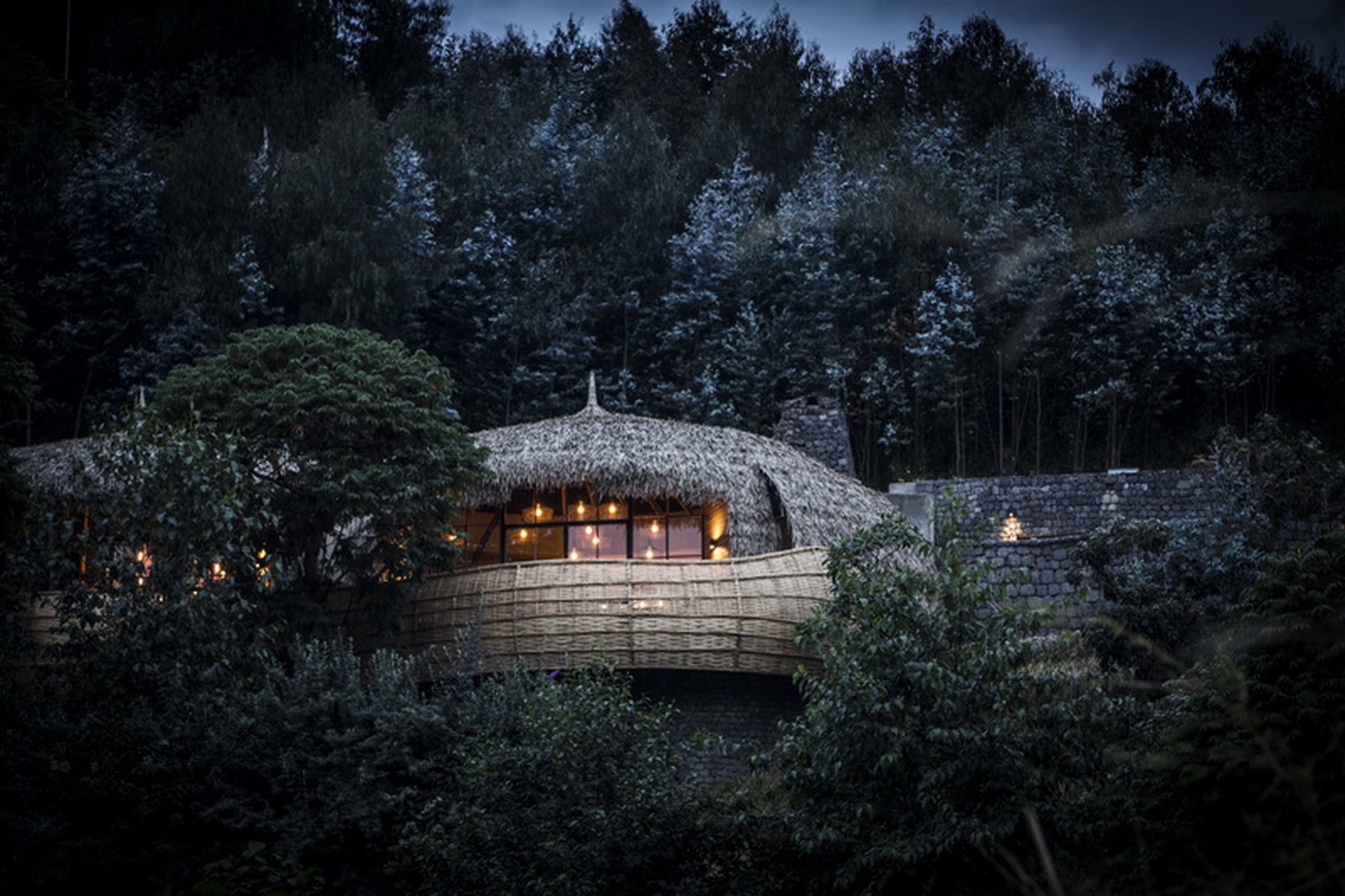 A warmly lit thatched lodge surrounded by forest in Rwanda at dusk.