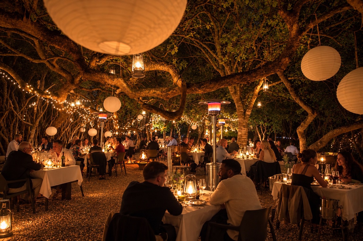 Guests dining beneath lanterns and trees at an outdoor evening gathering.