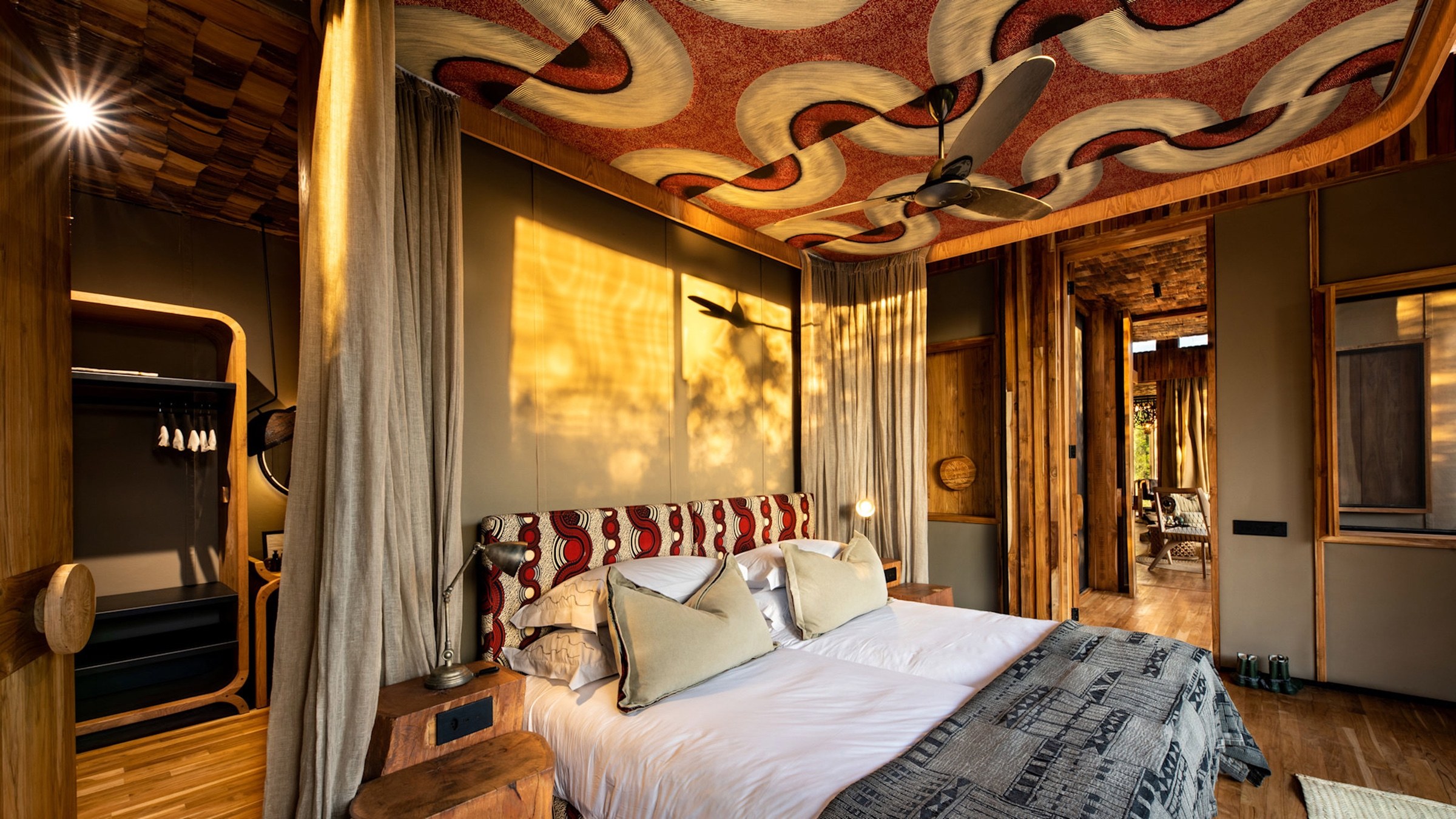 A safari lodge bedroom with timber finishes and a patterned ceiling in warm evening light.