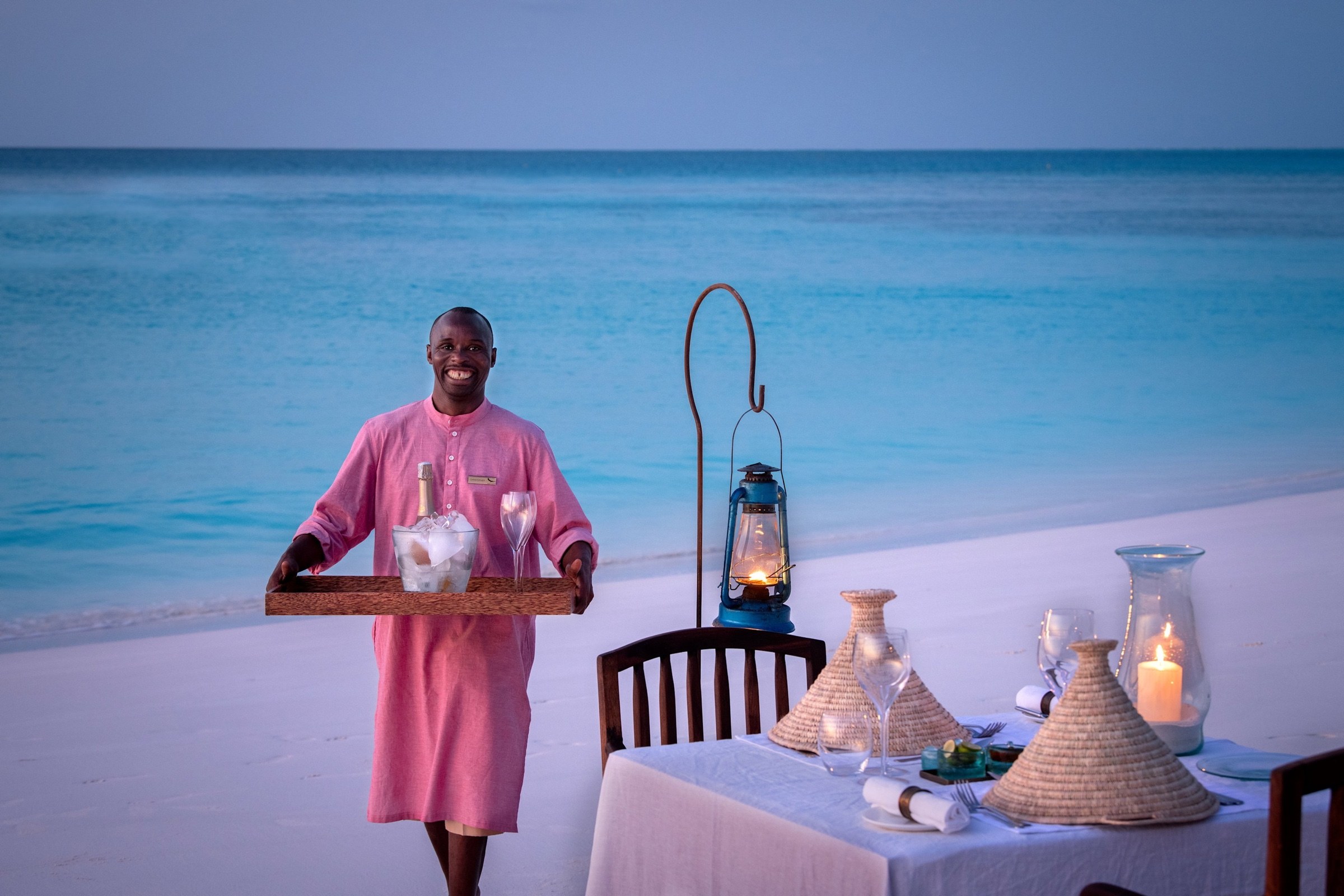 A host carrying drinks towards a candlelit dinner on a Zanzibar beach.