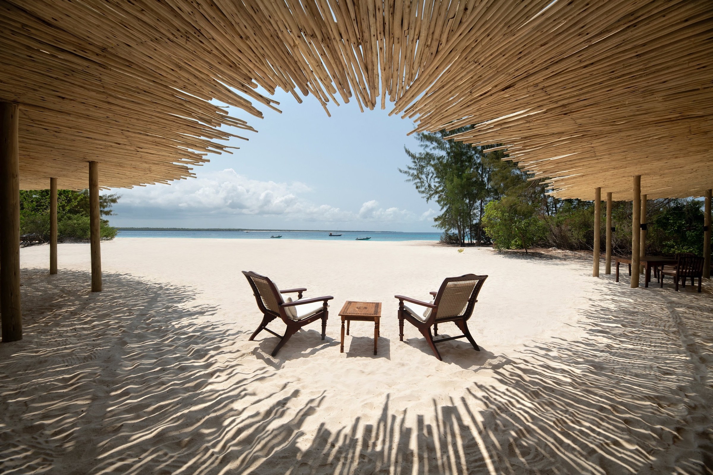 Two chairs beneath a woven beach canopy overlooking white sand and the ocean in Zanzibar.