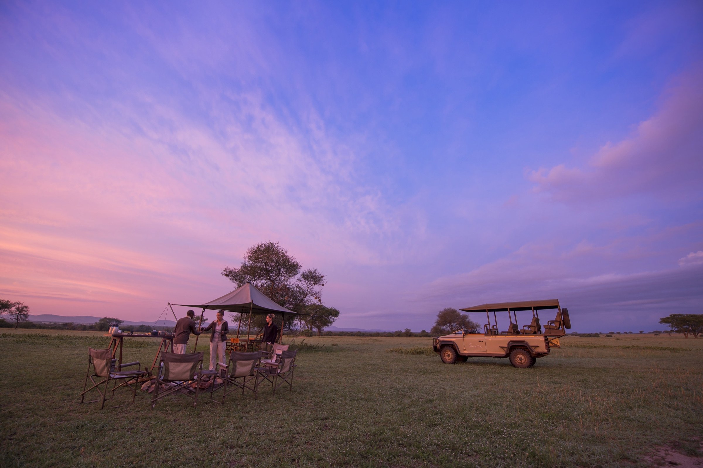 Guests and a camp host at a sundowner setup beside a safari vehicle under a pink sky.