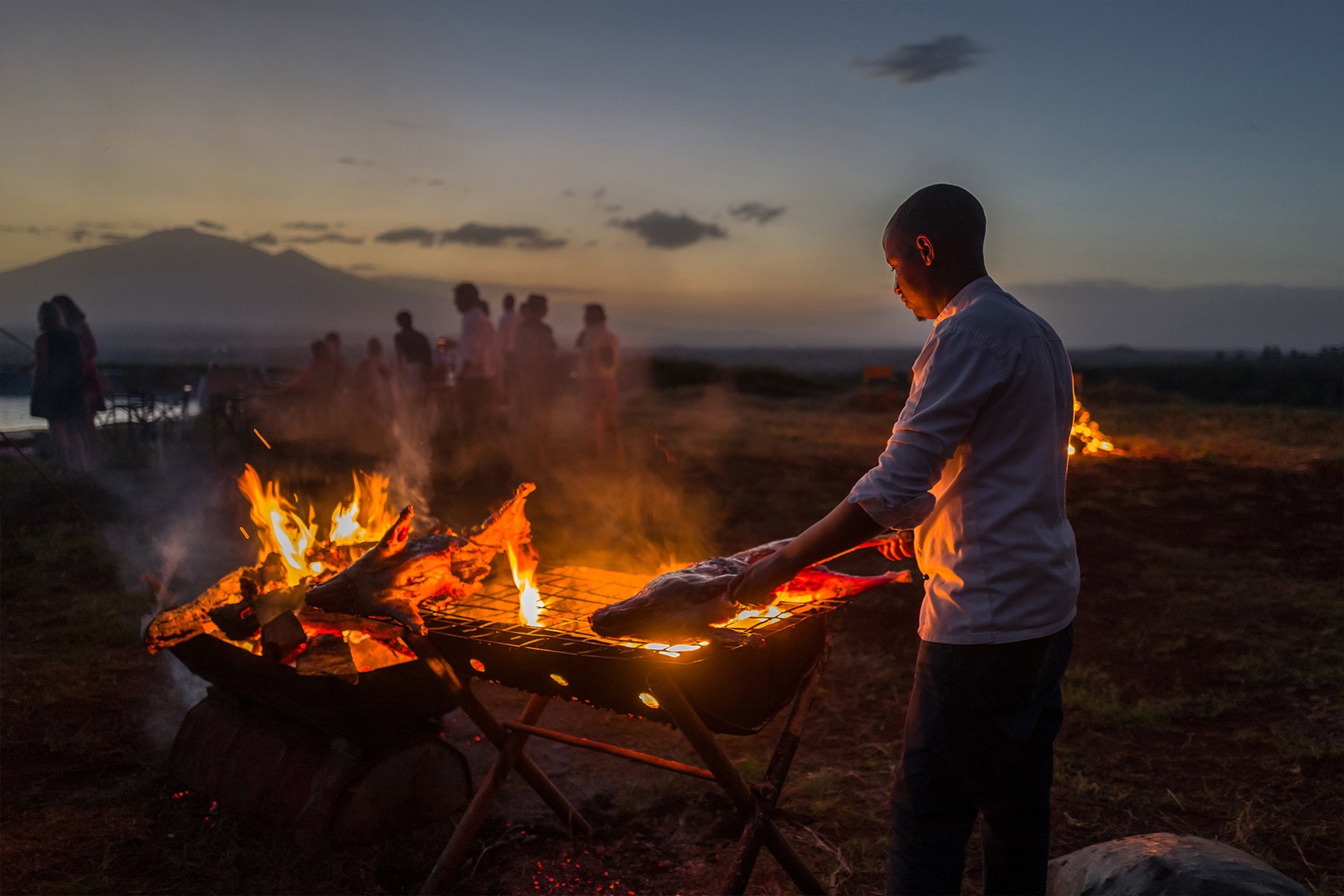 A chef cooking over an open fire with safari guests gathered at sunset.