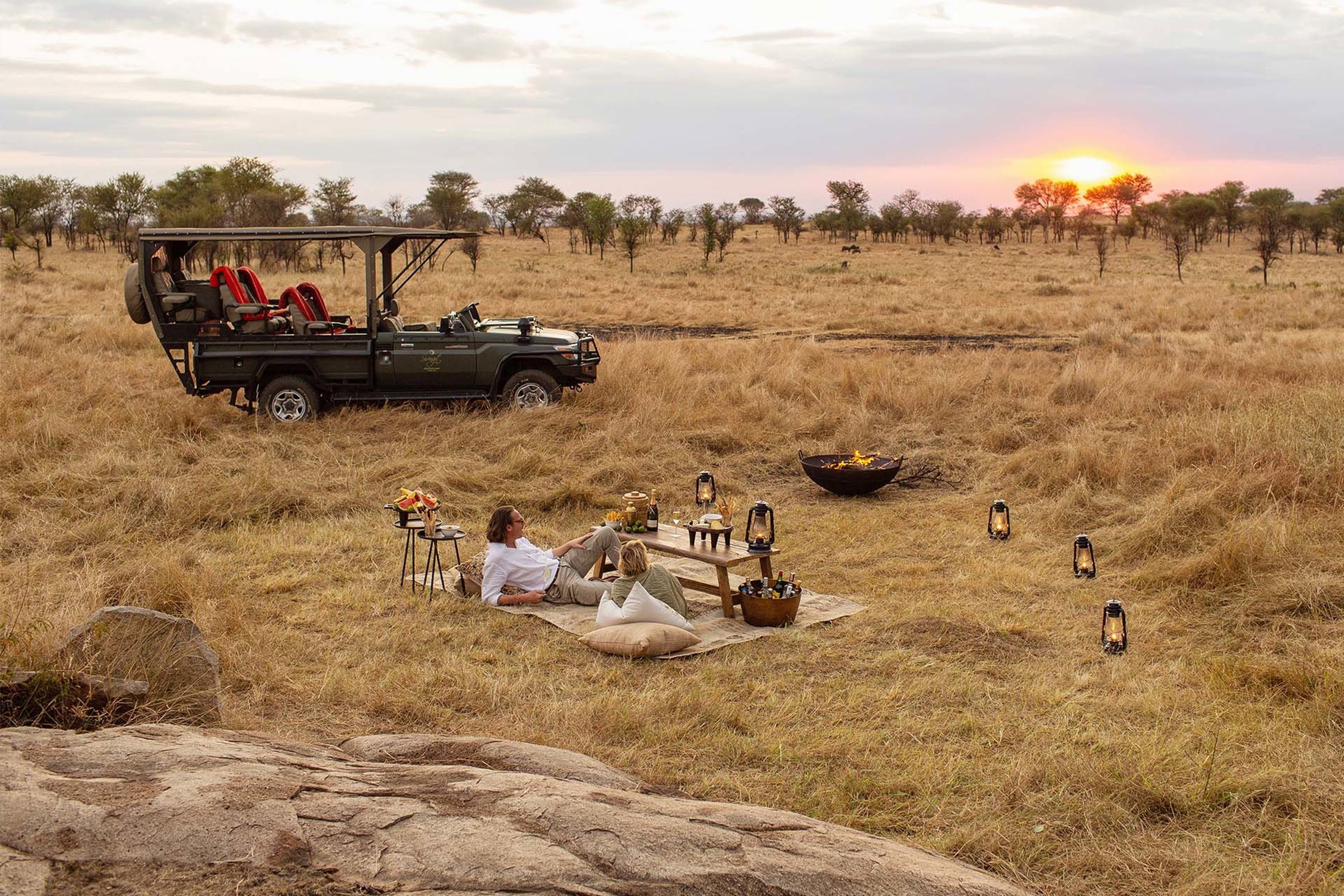 Two guests relaxing at a private sundowner picnic beside a safari vehicle in golden grass.