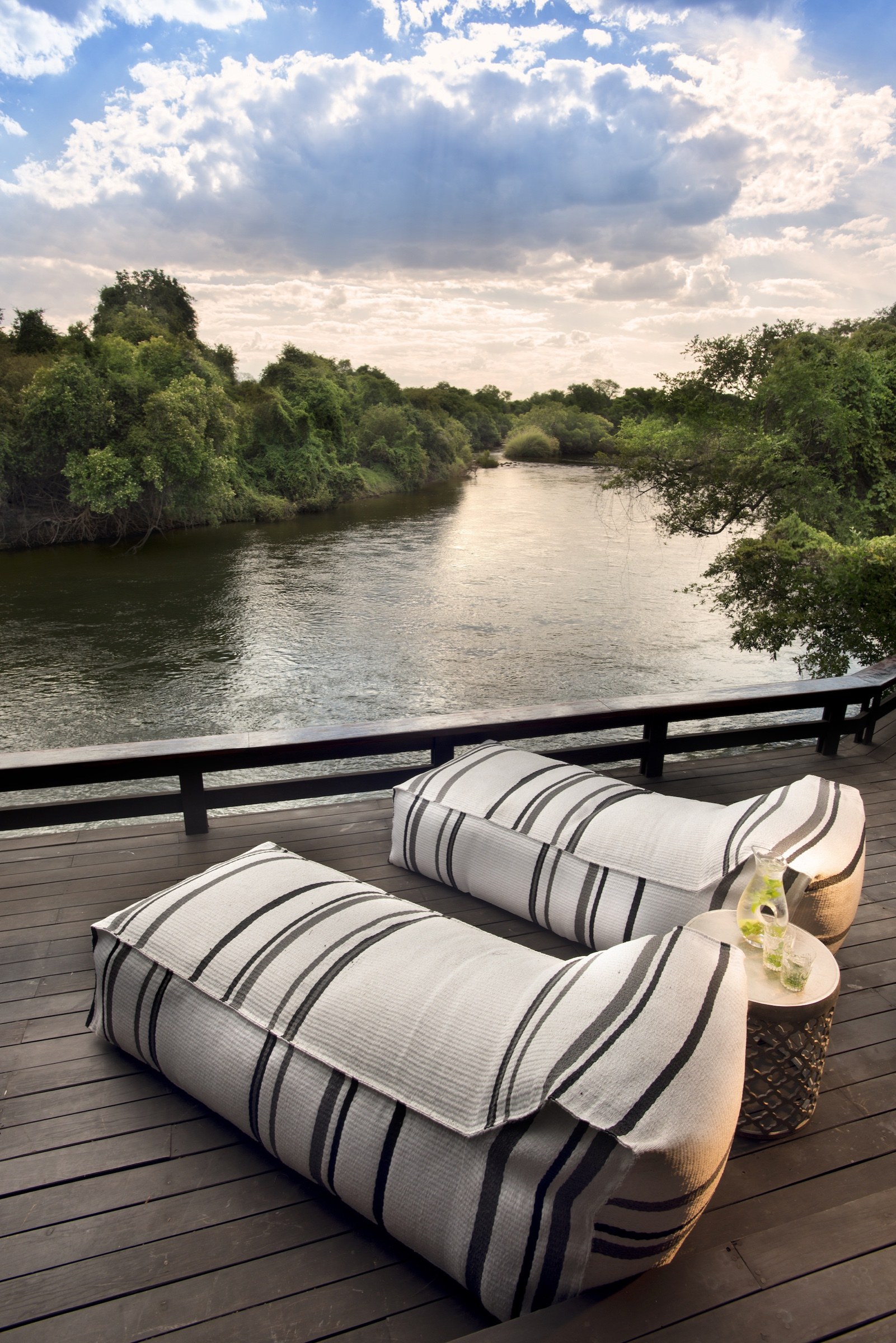 Cushioned seats and a small table on a wooden deck overlooking a tree-lined river
